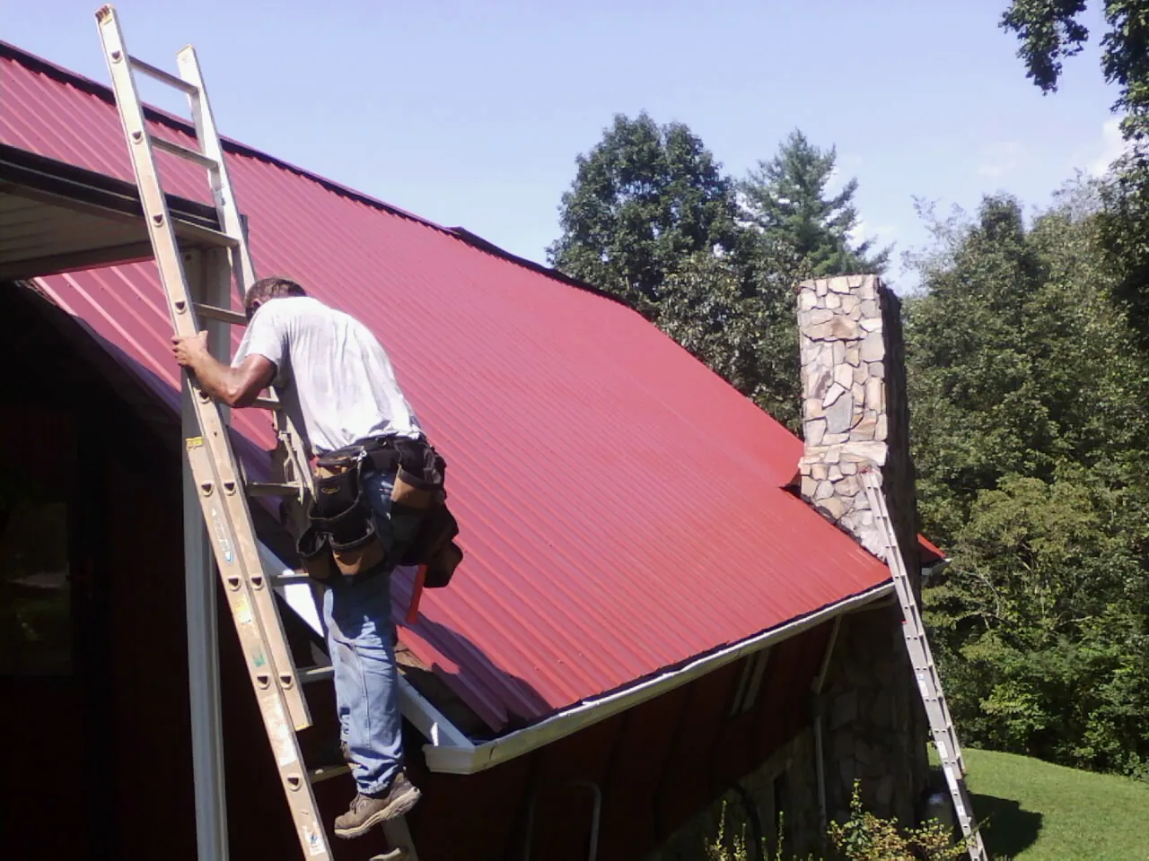 Roofer on a ladder beside a red metal roof and stone chimney — Soffit & Fascia Repair by Roofing Brothers in Wayzata